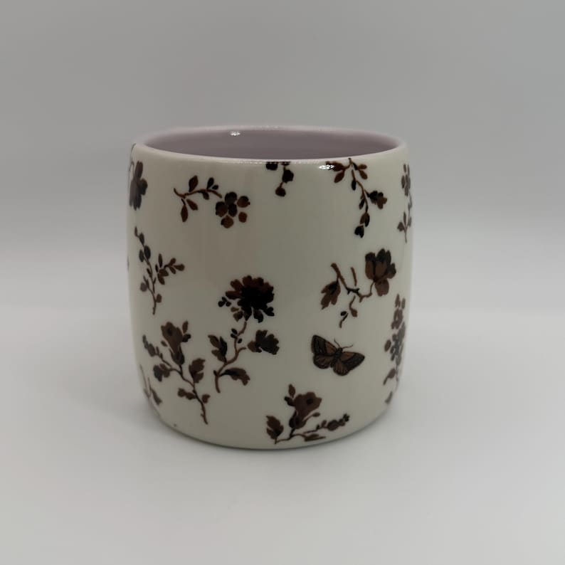May include: A ceramic tumbler with a cream-colored exterior adorned with brown floral and butterfly patterns. The interior rim is a soft pink. The tumbler is cylindrical and suitable for beverages.