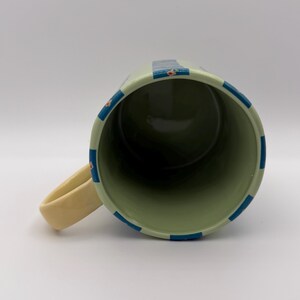 May include: A ceramic mug with a light green exterior, adorned with blue stripes and small floral accents. The interior is a darker green. The handle is a cream color. The mug is designed for hot or cold beverages.