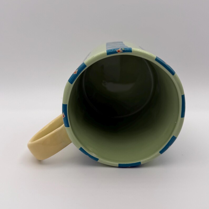 May include: A ceramic mug with a light green exterior, adorned with blue stripes and small floral accents. The interior is a darker green. The handle is a cream color. The mug is designed for hot or cold beverages.