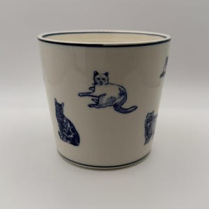 May include: A white ceramic tumbler with a navy blue rim and cat illustrations. The cats are in various poses and are also navy blue. The tumbler is cylindrical and has a smooth surface.