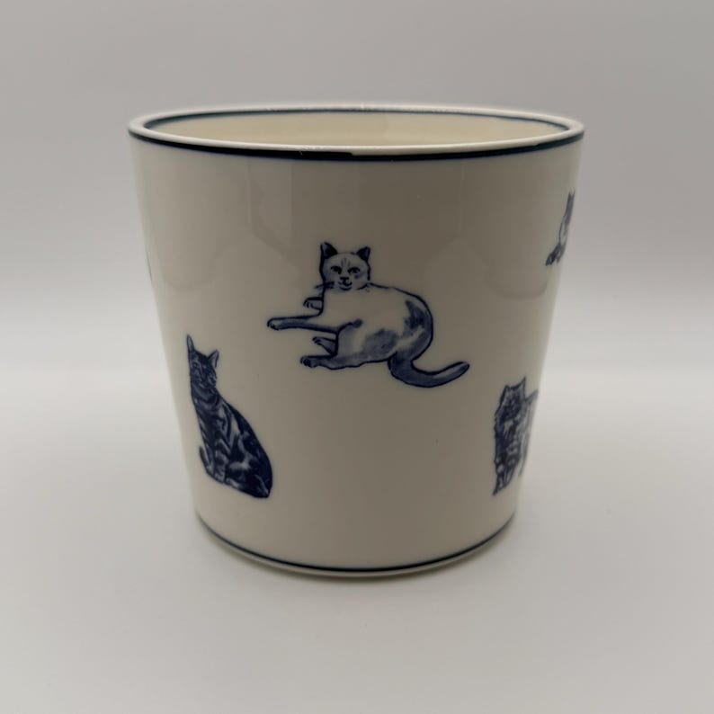 May include: A white ceramic tumbler with a navy blue rim and cat illustrations. The cats are in various poses and are also navy blue. The tumbler is cylindrical and has a smooth surface.