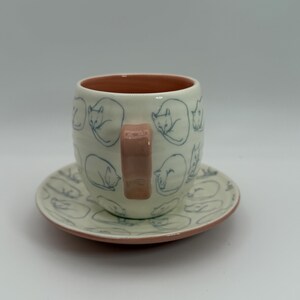 May include: A ceramic teacup and saucer set. The off-white cup and saucer feature a repeating pattern of sleeping cats in blue. The cup has a pink handle and interior, and the saucer has a pink rim. A charming cat-themed design.