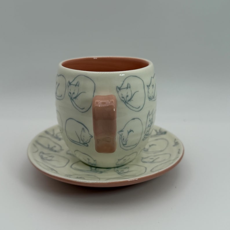 May include: A ceramic teacup and saucer set. The off-white cup and saucer feature a repeating pattern of sleeping cats in blue. The cup has a pink handle and interior, and the saucer has a pink rim. A charming cat-themed design.