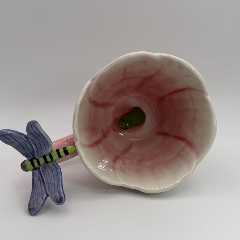 May include: A ceramic, flower-shaped bowl with a pink interior and a green center. A blue and green dragonfly is attached to the side of the bowl. The bowl has a white exterior and a pink stem.