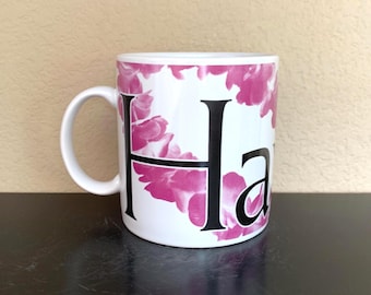May include: White ceramic mug with a pink floral design and the word "Ha" in black. The mug has a curved handle and is set against a neutral background.