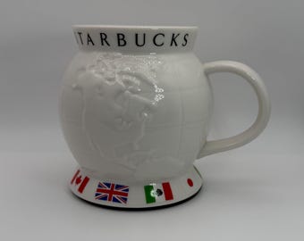 May include: White ceramic Starbucks mug with a globe design. The mug features the word "STARBUCKS" in black lettering and flags of different countries around the base. The mug has a rounded shape and a large handle.