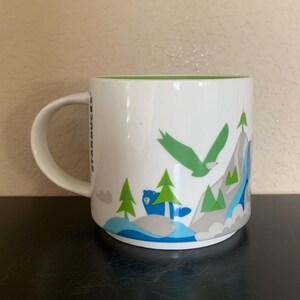 May include: White ceramic mug with a green interior and a handle. The mug features a mountain scene with a green eagle, blue bear, green trees, and gray mountains. The word "STARBUCKS" is printed vertically on the side.