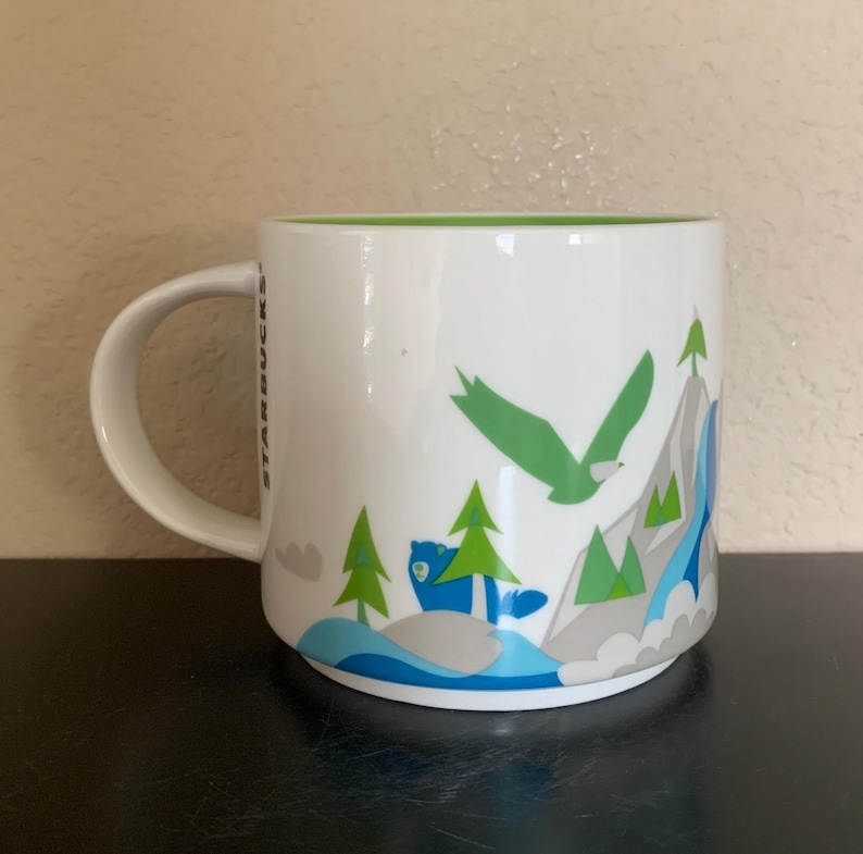 May include: White ceramic mug with a green interior and a handle. The mug features a mountain scene with a green eagle, blue bear, green trees, and gray mountains. The word "STARBUCKS" is printed vertically on the side.