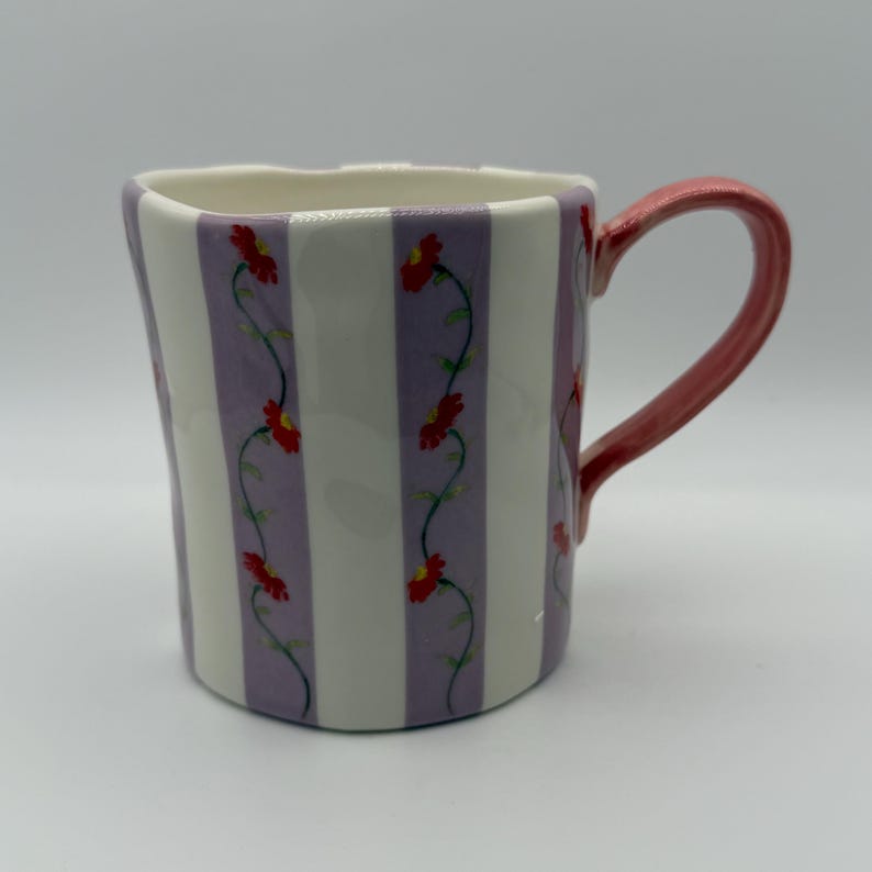 Pu&ograve; includere: Una tazza in ceramica dalla forma ondulata unica. La tazza presenta strisce verticali bianche e lavanda, con un motivo a vite floreale di fiori rossi e steli verdi. Il manico &egrave; di colore rosa pieno.