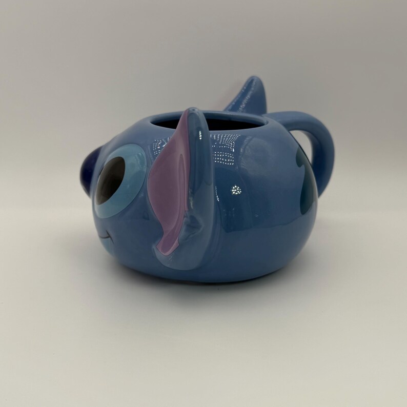 Puede incluir: Una taza de cer&aacute;mica azul con la forma del personaje Stitch. La taza presenta la cara de Stitch, con grandes ojos azules, orejas moradas y un asa peque&ntilde;a. La taza es brillante y tiene un acabado liso.