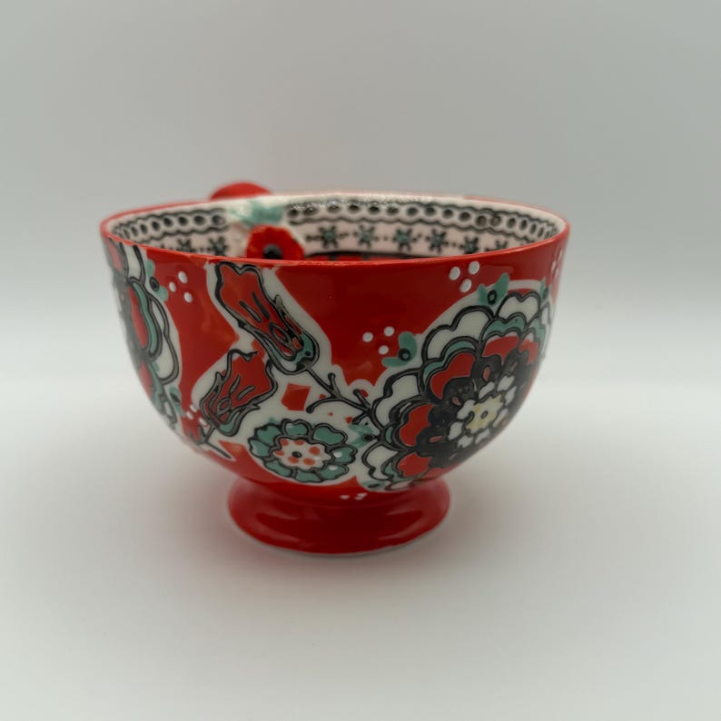 Może przedstawiać: Czerwona ceramiczna miska z kwiatowym wzorem w kolorze czarnym, białym i turkusowym. Miska ma podstawę i mały uchwyt. Obręcz jest ozdobiona czarno-białym wzorem.
