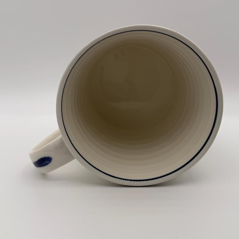 May include: White ceramic mug with a blue accent on the handle and a thin blue line around the rim. The interior has a series of concentric rings. The mug is empty and appears to be clean.