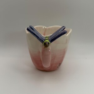 May include: A ceramic mug with a dragonfly handle. The mug is painted with a gradient of pink and white. The dragonfly has blue wings and a green and brown body. The mug is designed for beverages.