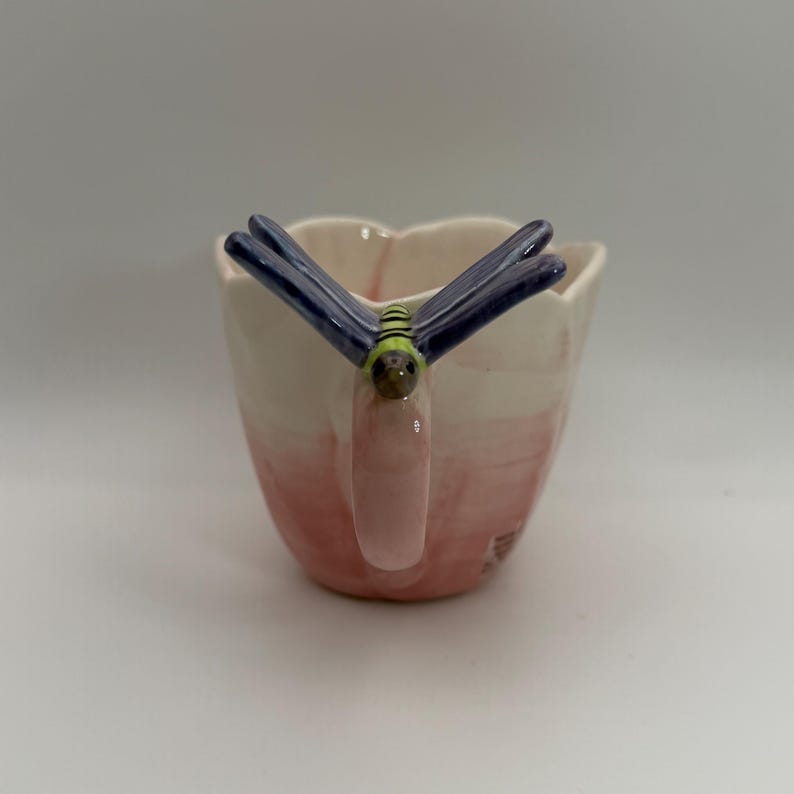 May include: A ceramic mug with a dragonfly handle. The mug is painted with a gradient of pink and white. The dragonfly has blue wings and a green and brown body. The mug is designed for beverages.