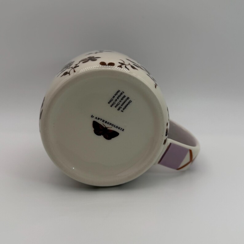 May include: A white ceramic mug with a floral pattern in brown and a lavender handle. The bottom of the mug features a butterfly graphic and the text "by ANTHROPOLOGIE". The mug is designed for hot beverages.