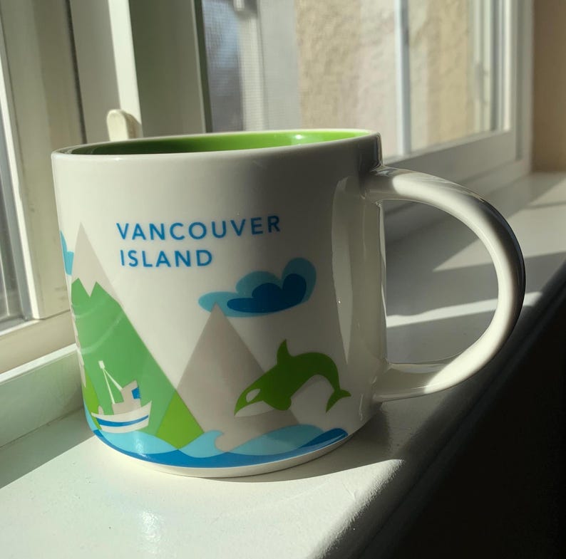 May include: White ceramic mug with a green interior and a curved handle. The mug features the text "VANCOUVER ISLAND" in blue, along with illustrations of mountains, a boat, a dolphin, and clouds in shades of green and blue.