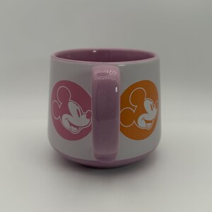 Puede incluir: Una taza de cer&aacute;mica blanca con interior y asa rosas. La taza presenta siluetas de Mickey Mouse en c&iacute;rculos rosas y naranjas en lados opuestos. El asa y el interior son de color rosa a juego.