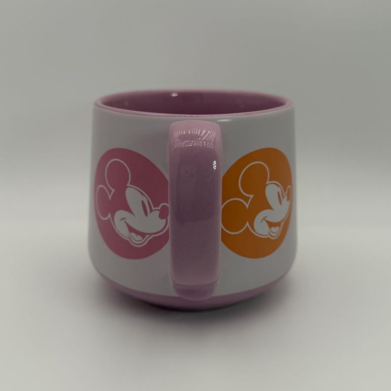 Puede incluir: Una taza de cer&aacute;mica blanca con interior y asa rosas. La taza presenta siluetas de Mickey Mouse en c&iacute;rculos rosas y naranjas en lados opuestos. El asa y el interior son de color rosa a juego.
