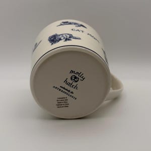 May include: Cream-colored ceramic mug with a black rim and handle. The mug features blue cat illustrations and the words "CAT MOM." The bottom of the mug has the text "molly hatch exclusively for ANTHROPOLOGIE."