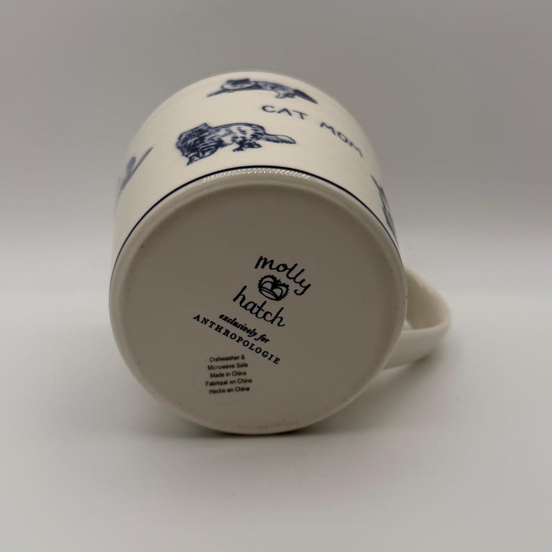 May include: Cream-colored ceramic mug with a black rim and handle. The mug features blue cat illustrations and the words "CAT MOM." The bottom of the mug has the text "molly hatch exclusively for ANTHROPOLOGIE."