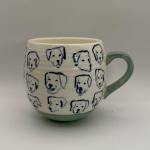 May include: A ceramic mug with a white base and a light green handle and bottom. The mug is decorated with blue line drawings of dog faces. The mug is a rounded shape.