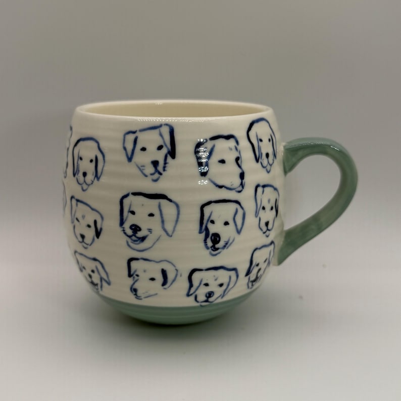 May include: A ceramic mug with a white base and a light green handle and bottom. The mug is decorated with blue line drawings of dog faces. The mug is a rounded shape.