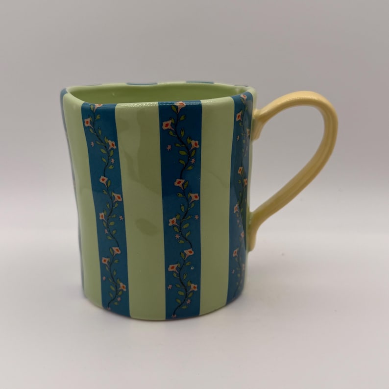 May include: A ceramic mug with a light green background and vertical blue stripes. The blue stripes are decorated with a floral pattern. The mug has a yellow handle.