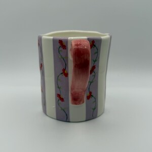 Pu&ograve; includere: Una tazza in ceramica dalla forma unica, con strisce verticali lavanda e bianche. La tazza &egrave; decorata con un motivo floreale di fiori rossi e tralci verdi. Il manico &egrave; di colore rosa pieno.