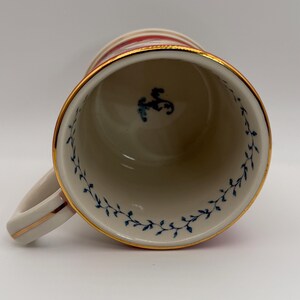 May include: A white ceramic mug with a gold rim and handle. The mug features red and gold stripes around the top and a blue floral design inside. A small blue emblem is visible inside the mug.