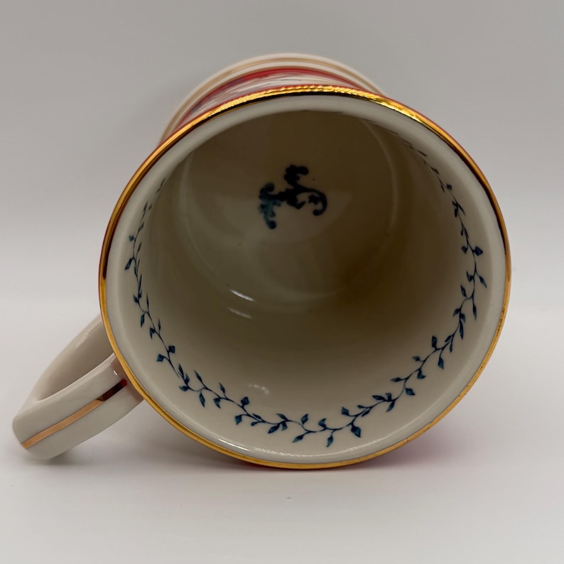 May include: A white ceramic mug with a gold rim and handle. The mug features red and gold stripes around the top and a blue floral design inside. A small blue emblem is visible inside the mug.
