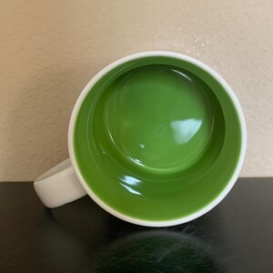May include: Overhead view of a white ceramic mug with a bright green interior. The mug has a white handle and a white rim. The mug is sitting on a black surface.