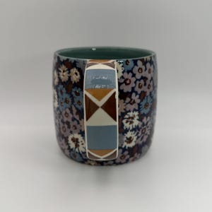 Puede incluir: Una taza de cer&aacute;mica con un estampado floral en tonos azules, morados y blancos. El asa presenta un dise&ntilde;o geom&eacute;trico con formas marrones, azules y blancas. El interior de la taza es de color verde liso.