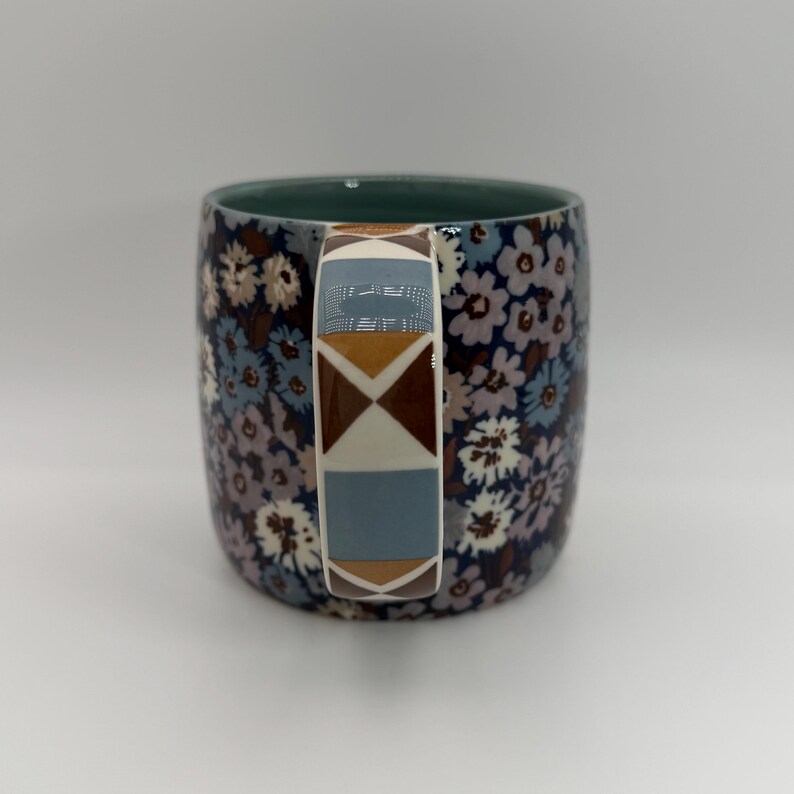 Puede incluir: Una taza de cer&aacute;mica con un estampado floral en tonos azules, morados y blancos. El asa presenta un dise&ntilde;o geom&eacute;trico con formas marrones, azules y blancas. El interior de la taza es de color verde liso.