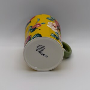 K&ouml;nnte beinhalten: Gelbe Blumenvase mit gr&uuml;nem Griff. Die Tasse hat ein Muster aus rosa und wei&szlig;en Rosen und gr&uuml;nen Bl&auml;ttern auf gelbem Hintergrund. Der Boden der Tasse ist wei&szlig; mit dem Markennamen "Maeve".