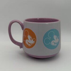 Puede incluir: Una taza de cer&aacute;mica con exterior gris claro, asa morada e interior morado. La taza presenta dos dise&ntilde;os de Mickey Mouse en c&iacute;rculos naranja y azul. La base de la taza tambi&eacute;n es morada.