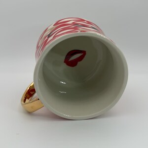 Pode incluir: Uma caneca de cer&acirc;mica branca com uma al&ccedil;a dourada. O exterior da caneca apresenta um padr&atilde;o de l&aacute;bios vermelhos e brancos. Dentro da caneca, um gr&aacute;fico de l&aacute;bios vermelhos &eacute; vis&iacute;vel. A caneca &eacute; projetada para bebidas quentes.
