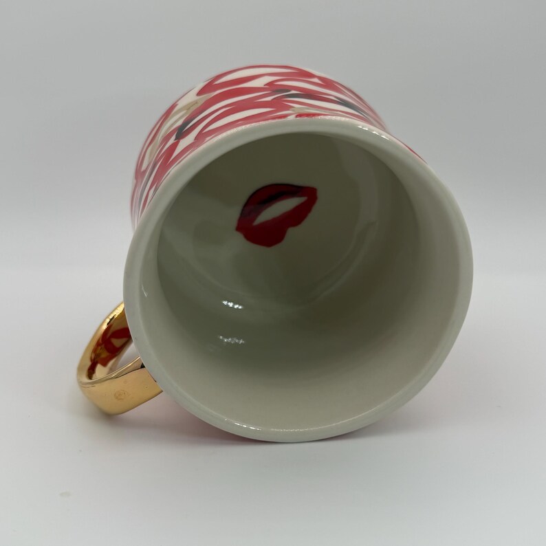 Pode incluir: Uma caneca de cer&acirc;mica branca com uma al&ccedil;a dourada. O exterior da caneca apresenta um padr&atilde;o de l&aacute;bios vermelhos e brancos. Dentro da caneca, um gr&aacute;fico de l&aacute;bios vermelhos &eacute; vis&iacute;vel. A caneca &eacute; projetada para bebidas quentes.