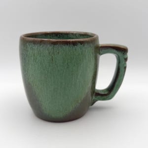 May include: A ceramic mug featuring a two-tone glaze with a dark brown base that transitions into a mottled sage green finish on the upper body and handle. The mug has a rounded, cylindrical shape with a sturdy, thick-walled handle.