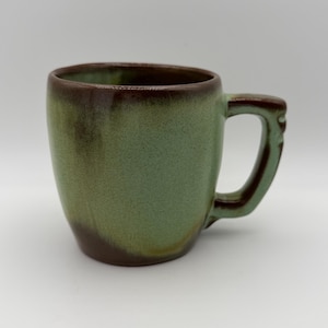 May include: A ceramic mug featuring a rounded body with a reactive glaze finish in shades of sage green and deep brown. The mug has a thick, sturdy handle and a smooth, curved rim, showcasing a blend of earthy tones across the exterior surface.