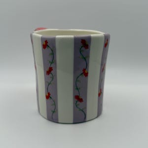 Pu&ograve; includere: Una tazza in ceramica con un motivo a righe bianche e lavanda. Fiori rossi con viti verdi decorano le strisce lavanda. La tazza ha un piccolo manico rosa.