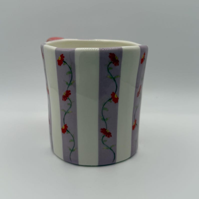 Pu&ograve; includere: Una tazza in ceramica con un motivo a righe bianche e lavanda. Fiori rossi con viti verdi decorano le strisce lavanda. La tazza ha un piccolo manico rosa.