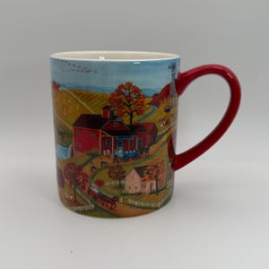 May include: A ceramic mug with a rural autumn scene. The mug features a red handle and a detailed illustration of a farm with red barns, a windmill, and a horse-drawn carriage. The background shows rolling hills and a blue sky.