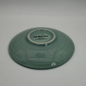 May include: A teal-colored ceramic dish with a slightly textured surface. The underside features the text "Leah Reena Goren" and "exclusively for ANTHROPOLOGIE" in black lettering. The dish has a shallow, rounded shape.