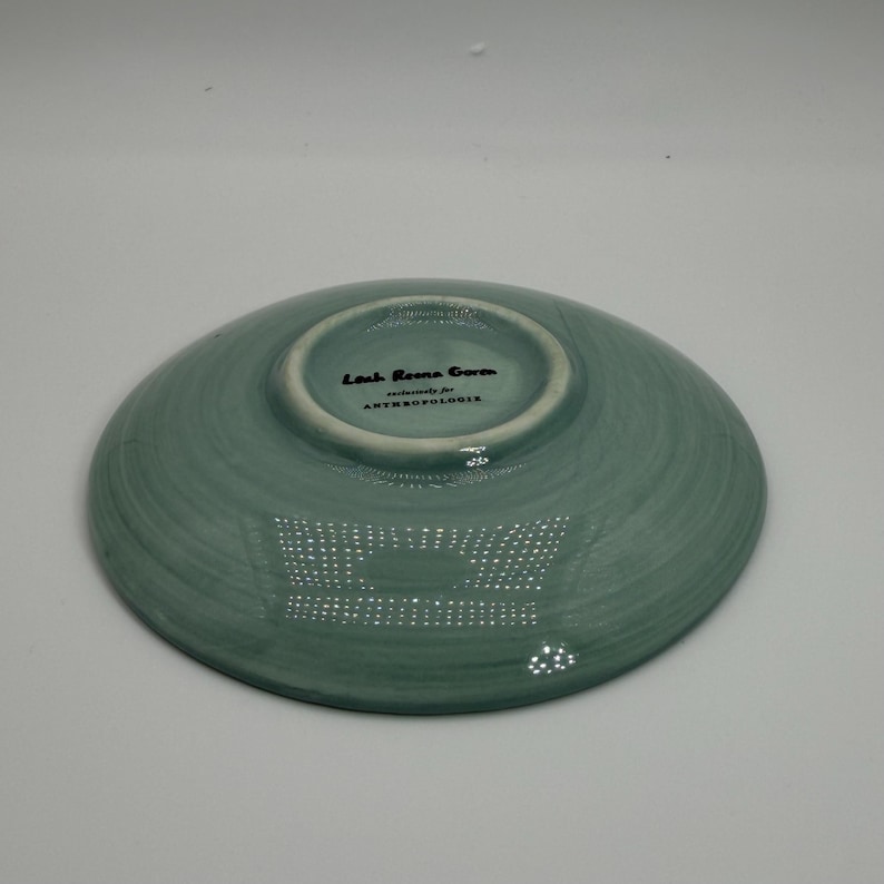May include: A teal-colored ceramic dish with a slightly textured surface. The underside features the text "Leah Reena Goren" and "exclusively for ANTHROPOLOGIE" in black lettering. The dish has a shallow, rounded shape.