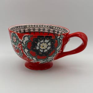 Może przedstawiać: Czerwony ceramiczny kubek do herbaty z dużym uchwytem i kwiatowym wzorem w kolorze czarnym, białym i turkusowym. Obręcz ma czarno-biały wz&oacute;r w kropki. Kubek stoi na małej, okrągłej podstawie.