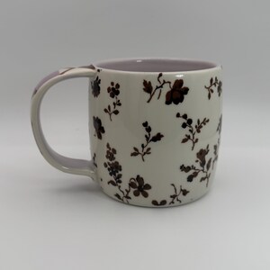 May include: Cream-colored ceramic mug with a brown floral pattern. The mug has a lavender interior and handle. The floral design includes various flower and leaf shapes. The handle has a lavender accent. A decorative kitchenware item.