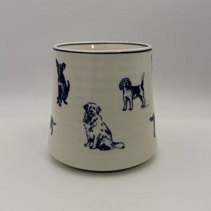 K&ouml;nnte beinhalten: Cremefarbene Keramiktasse mit dunkelblauen Hundeillustrationen. Die Hunde sind in verschiedenen Posen dargestellt. Die Tasse hat einen dunkelblauen Rand und eine leicht ausgestellte Form. H&ouml;he ca. 10 cm.
