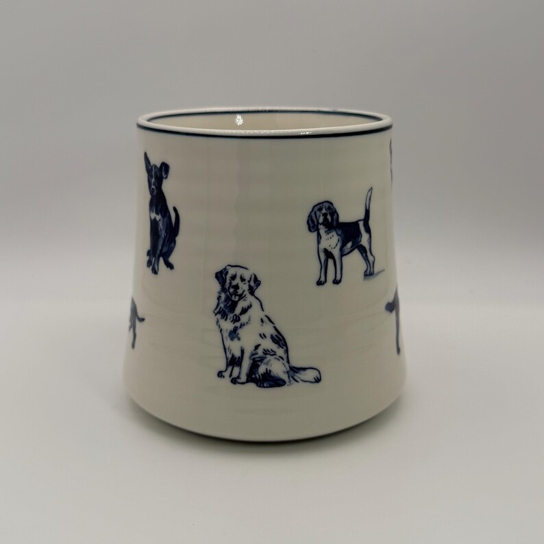 K&ouml;nnte beinhalten: Cremefarbene Keramiktasse mit dunkelblauen Hundeillustrationen. Die Hunde sind in verschiedenen Posen dargestellt. Die Tasse hat einen dunkelblauen Rand und eine leicht ausgestellte Form. H&ouml;he ca. 10 cm.
