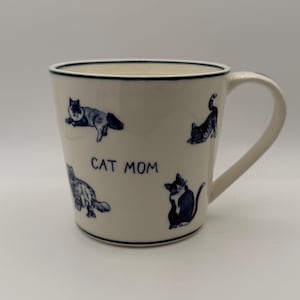 May include: White ceramic mug with a blue rim and handle. The mug features blue illustrations of cats in various poses and the text "CAT MOM". A charming gift for cat lovers.