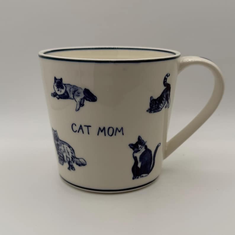 May include: White ceramic mug with a blue rim and handle. The mug features blue illustrations of cats in various poses and the text "CAT MOM". A charming gift for cat lovers.